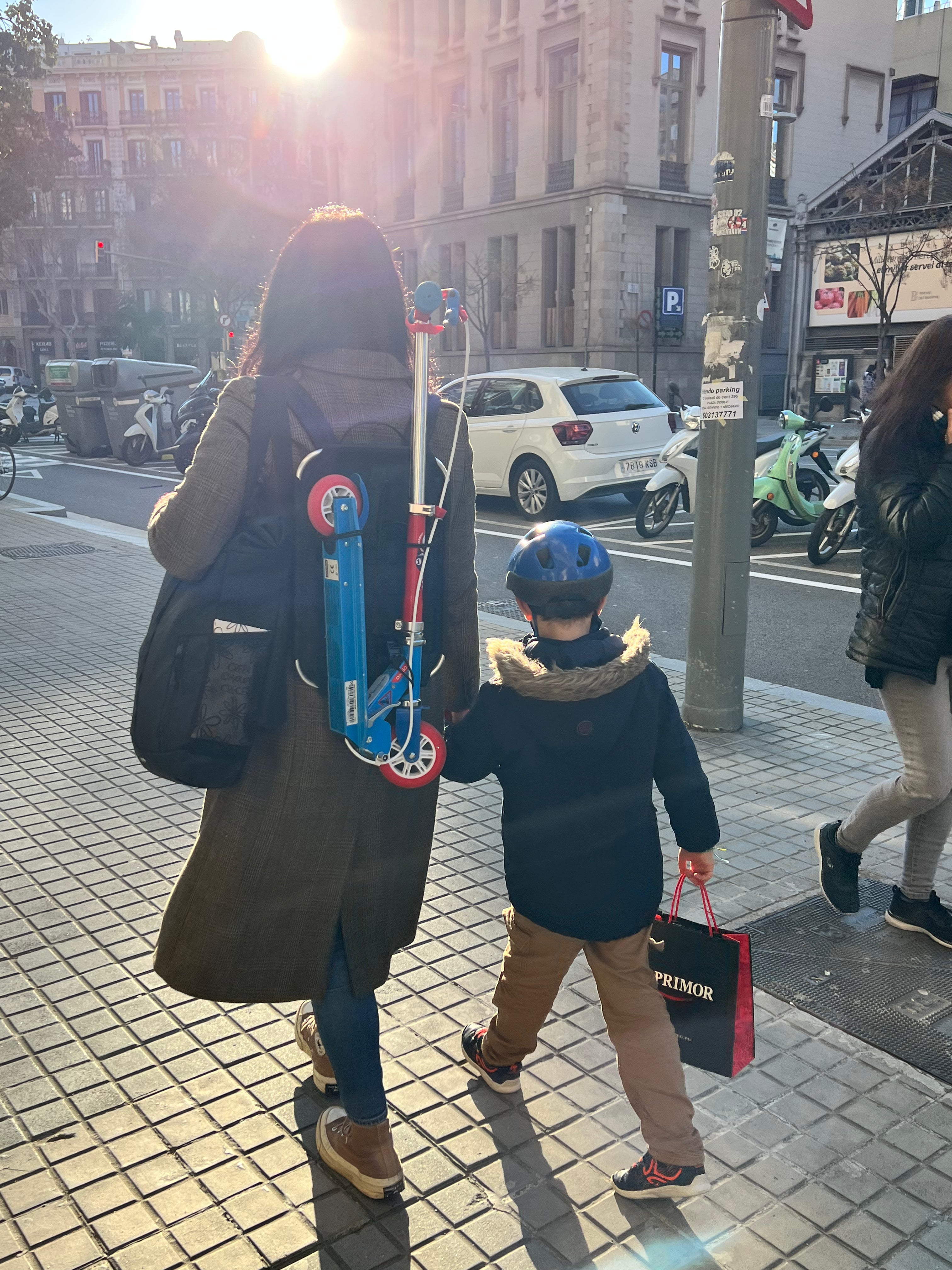 Madre caminando por una calle de Barcelona con el patinete de su hijo sujeto a la espalda, llevando al niño de la mano