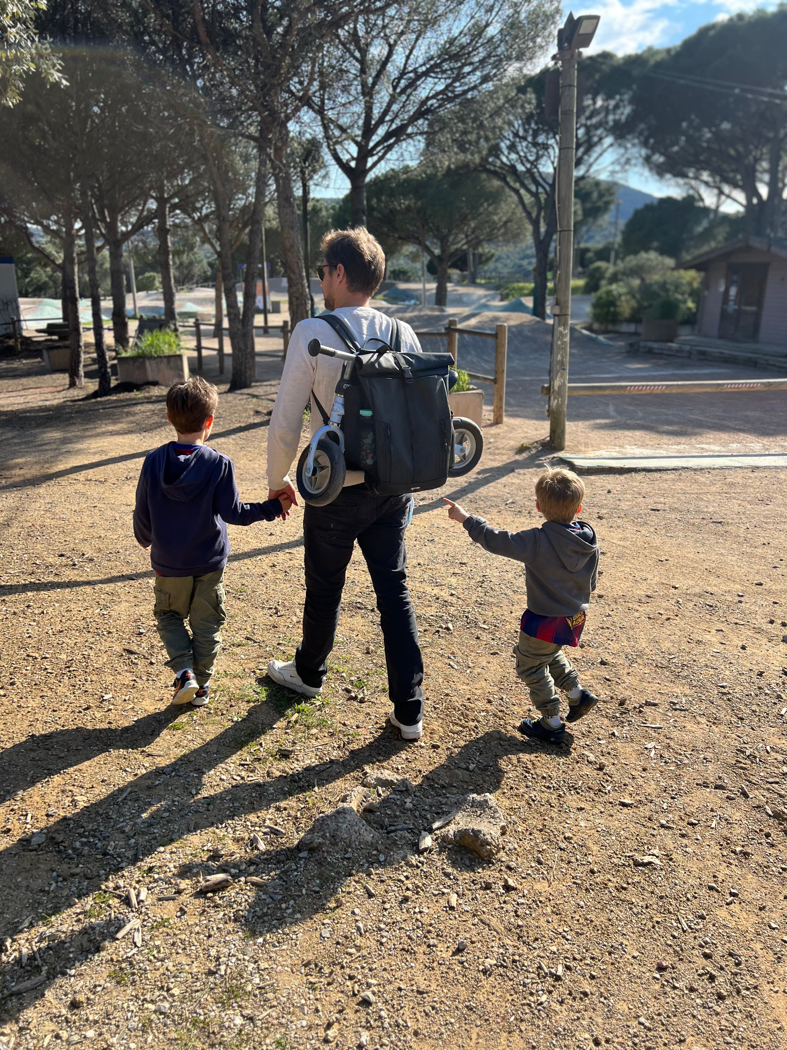 Padre de espaldas llevando el patinete de su hijo sujeto al BackPanel Iziride, paseando de la mano de sus dos niños por un camino de tierra